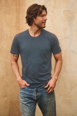 A man with medium-length brown hair wears the Velvet by Graham & Spencer HOWARD TEE in blue Pima jersey and jeans, standing against a textured beige wall with hands in pockets, evoking a subtle vintage vibe.