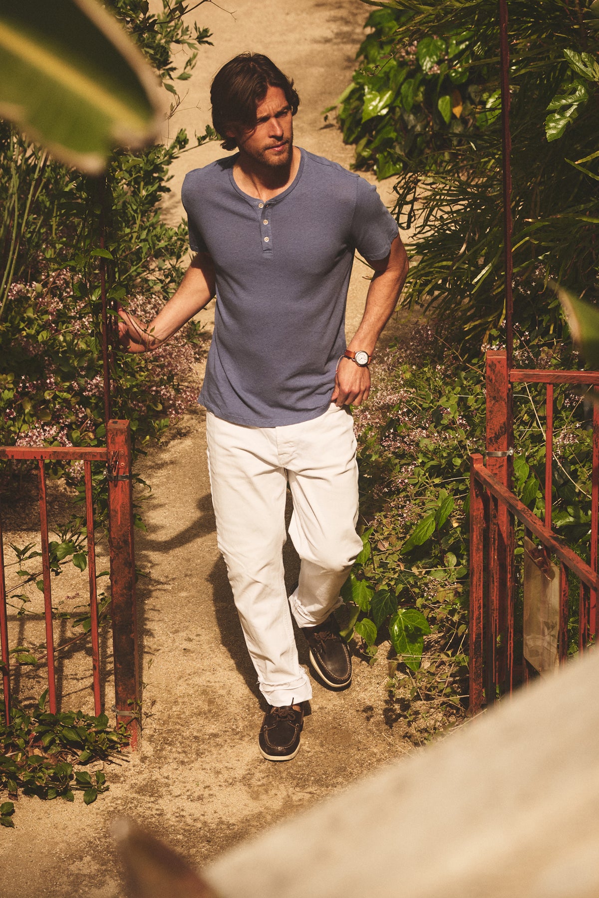 A man in the LIONEL LINEN COTTON HENLEY by Velvet by Graham & Spencer, paired with white pants and dark shoes, walks along a garden path bordered by green plants and rust-colored railings.-39152701112513