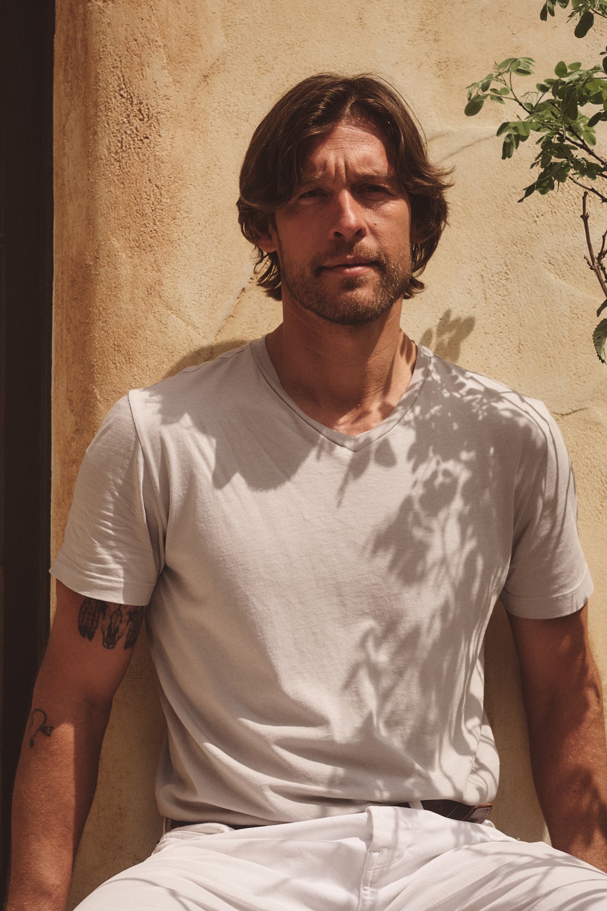 A man with medium-length hair and a beard sits in sunlight against a textured beige wall, wearing the Velvet by Graham & Spencer SAMSEN TEE in light gray and white pants. A plant's shadow adds depth to his effortless look.
