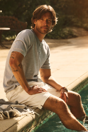 A man in the VITO HENLEY by Velvet by Graham & Spencer and white shorts sits on the pool's edge with his legs in the water, a towel at his side.