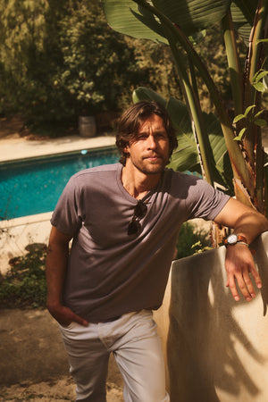 Wearing the ZANDER TEE by Velvet by Graham & Spencer and relaxed-fit white pants, a man leans against a concrete wall near a pool, sunglasses on his shirt and greenery behind him—an effortlessly wearable look.