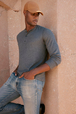 A man in jeans and a hat leaning against a wall, showcasing the Velvet by Graham & Spencer's ANTHONY THERMAL HENLEY's classic waffle texture.