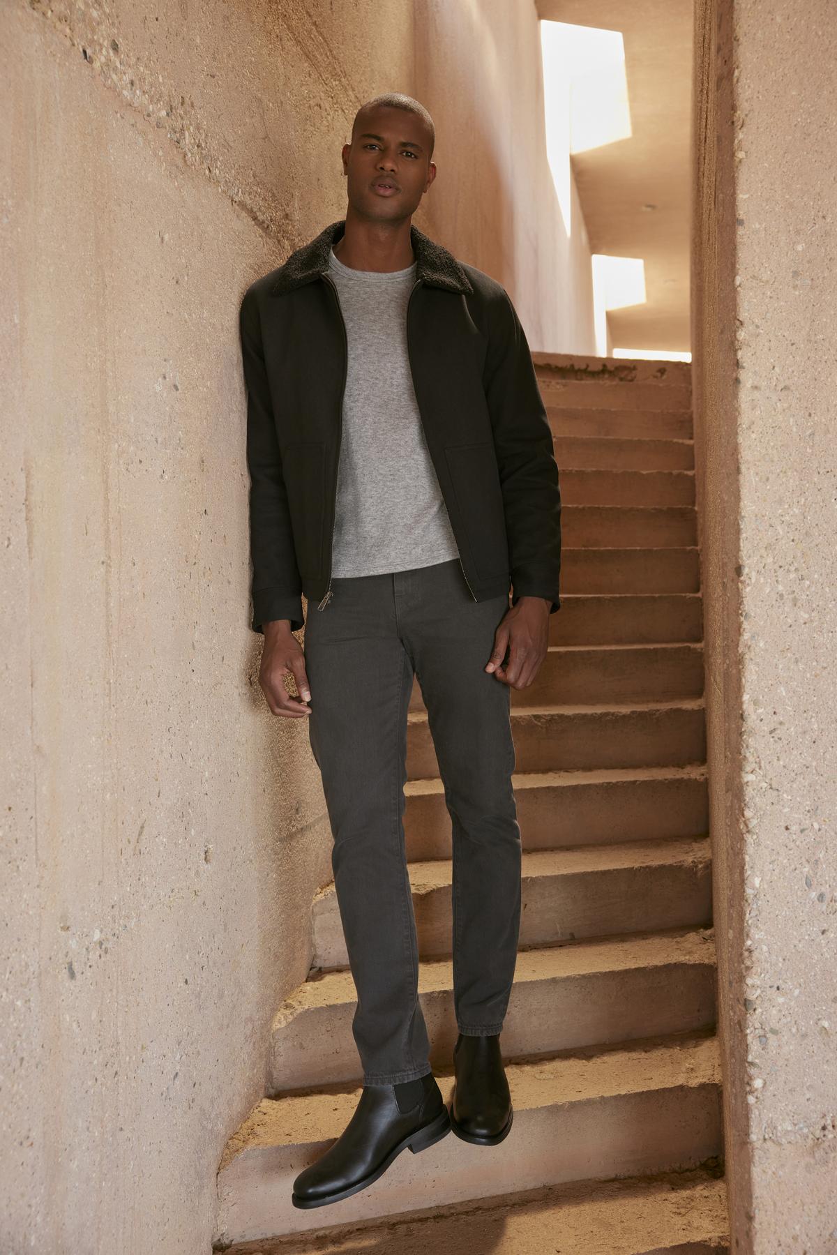 A person stands on stairs next to a wall, wearing a Velvet by Graham & Spencer CASH JACKET with a faux sherpa collar, gray shirt, gray pants, and black boots. The background is a concrete structure with rectangular light openings above.