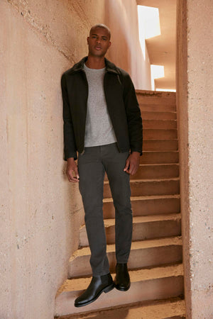 A person stands on stairs next to a wall, wearing a Velvet by Graham & Spencer CASH JACKET with a faux sherpa collar, gray shirt, gray pants, and black boots. The background is a concrete structure with rectangular light openings above.