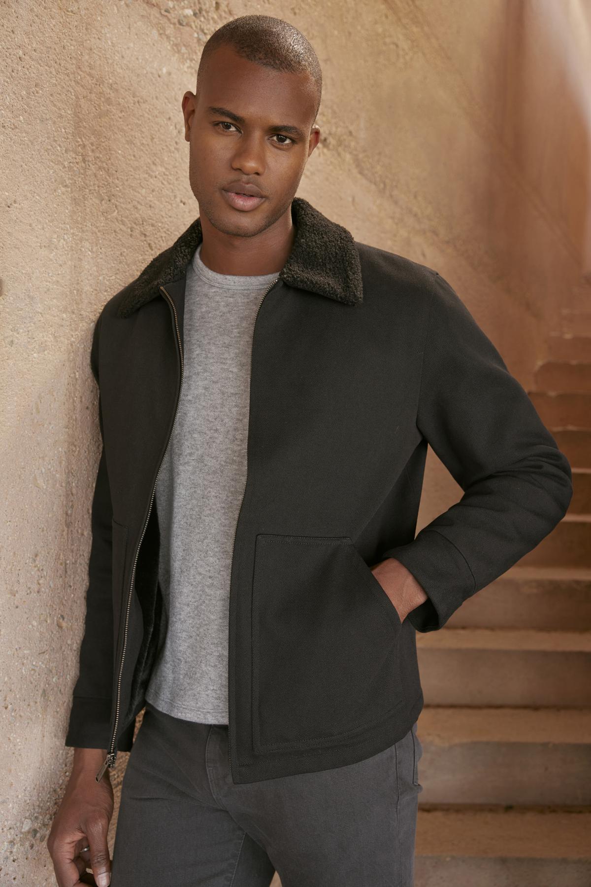 A person wearing a black Velvet by Graham & Spencer CASH JACKET with a faux sherpa collar and gray shirt stands by an outdoor staircase, looking at the camera.