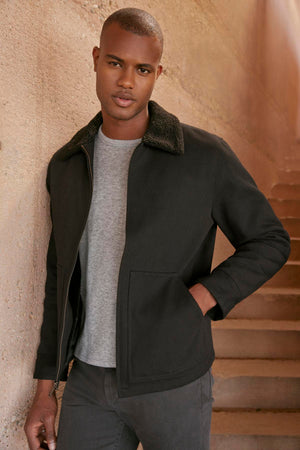 A person wearing a black Velvet by Graham & Spencer CASH JACKET with a faux sherpa collar and gray shirt stands by an outdoor staircase, looking at the camera.
