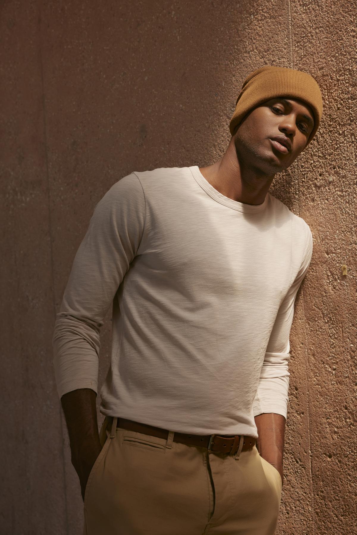 A person stands against a textured wall, wearing a light beige Velvet by Graham & Spencer KAI TEE with a classic crew neckline, camel-colored pants, and a matching brown beanie.-35547537965249