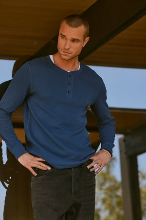 A man wearing the DEX THERMAL HENLEY from Velvet by Graham & Spencer with a 3-button placket and dark jeans stands under a shaded structure, looking to his side with his hands in his pockets. The cotton thermal knit adds an extra touch of warmth, making it perfect for layering on cooler days.