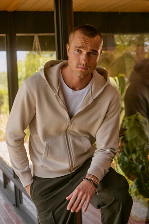 A person wearing a REN HOODIE by Velvet by Graham & Spencer along with dark pants stands indoors near a glass window, one hand casually tucked in their pocket.