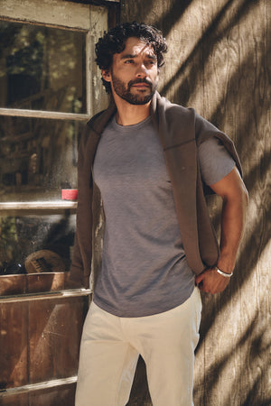 A man stands outside by a wooden wall and glass door, wearing the AMARO TEE by Velvet by Graham & Spencer—a gray Peruvian cotton crew neck—paired with beige pants and a brown sweater draped over his shoulders.
