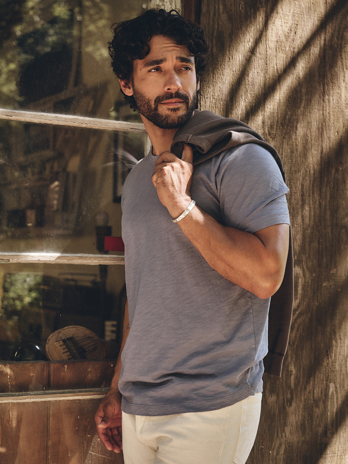 A man with curly hair and a beard stands outdoors, holding a jacket over his shoulder, wearing the AMARO TEE by Velvet by Graham & Spencer in light blue and beige pants.-40134803882177