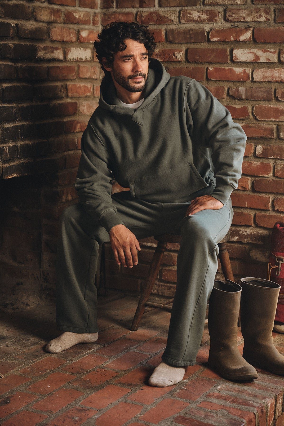 Wearing the KAZ HOODIE by Velvet by Graham & Spencer, a man sits indoors on a wooden chair against a brick wall, embodying everyday comfort and style.