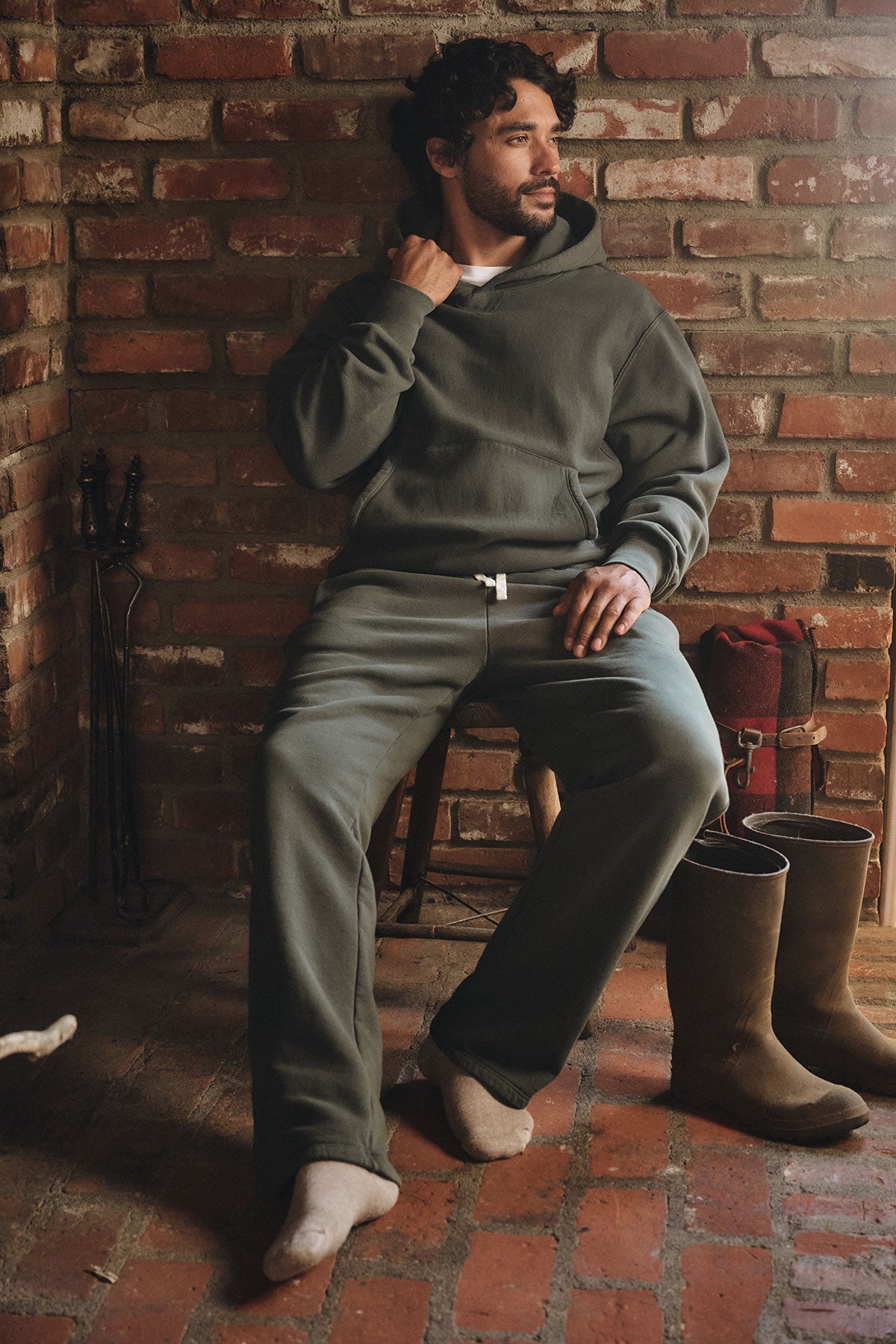 A man sits on a chair in a rustic brick room, wearing the Velvet by Graham & Spencer HUDSON SWEATPANT—a relaxed-fit cotton fleece piece—paired with a dark green hoodie, boots, and a red bag for cozy everyday style.