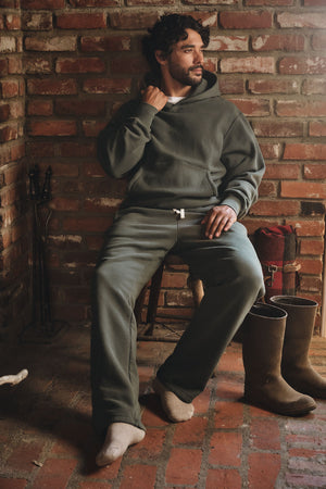 A man sits on a chair in a rustic brick room, wearing the Velvet by Graham & Spencer HUDSON SWEATPANT—a relaxed-fit cotton fleece piece—paired with a dark green hoodie, boots, and a red bag for cozy everyday style.