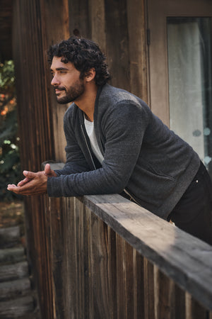 A man with dark hair and a beard leans on a wooden railing outside a rustic building, wearing the MILES JACKET by Velvet by Graham & Spencer—a gray knit shell with sherpa lining—over a white t-shirt and dark pants.
