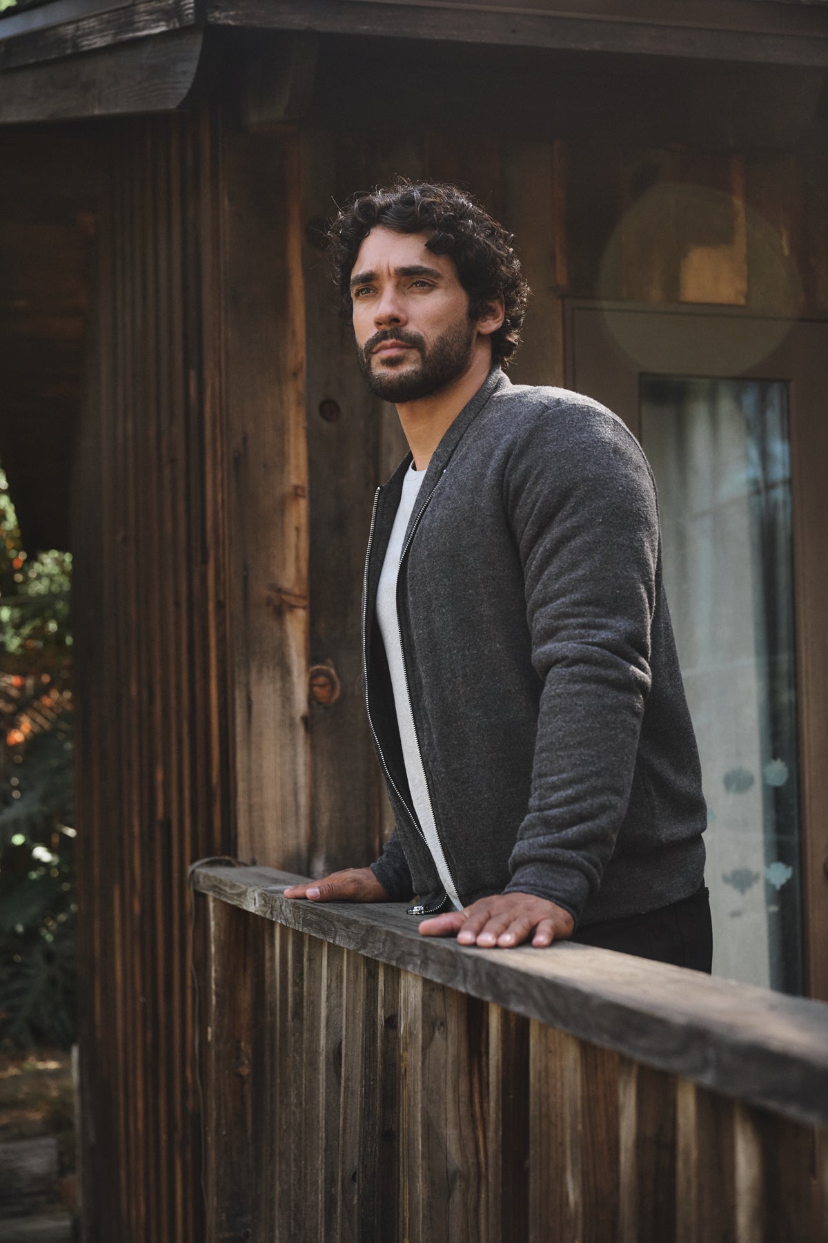A man with curly hair and a beard stands on a wooden porch, gazing into the distance while wearing the cozy MILES JACKET with soft sherpa lining by Velvet by Graham & Spencer.