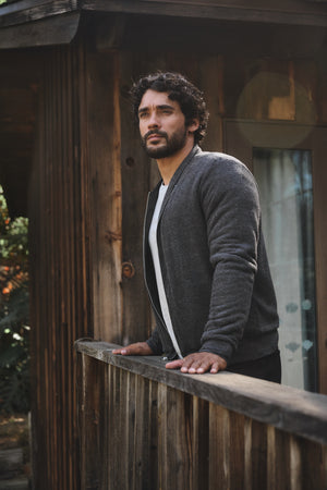 A man with curly hair and a beard stands on a wooden porch, gazing into the distance while wearing the cozy MILES JACKET with soft sherpa lining by Velvet by Graham & Spencer.