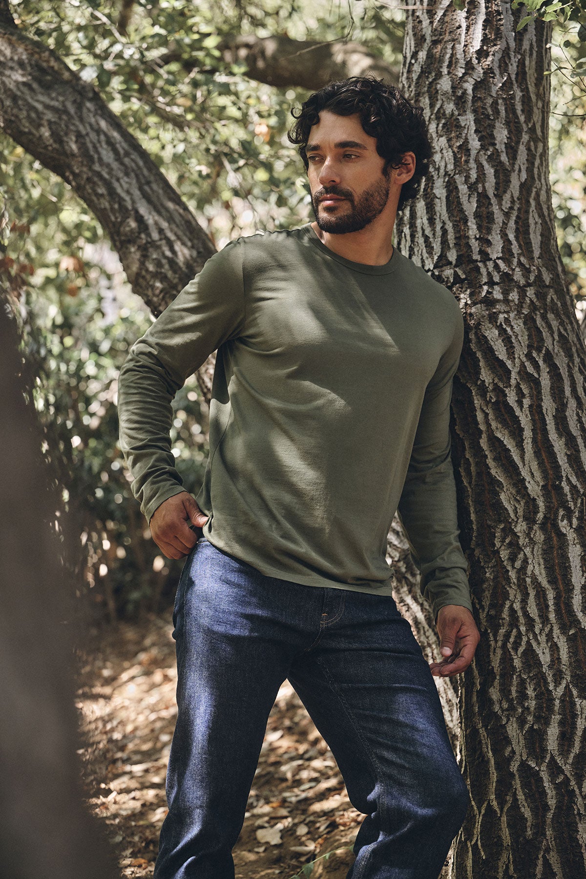 A man wearing the ROWLAND TEE by Velvet by Graham & Spencer in olive green, paired with blue jeans, stands outdoors leaning on a tree trunk against a wooded backdrop.-40147852656833
