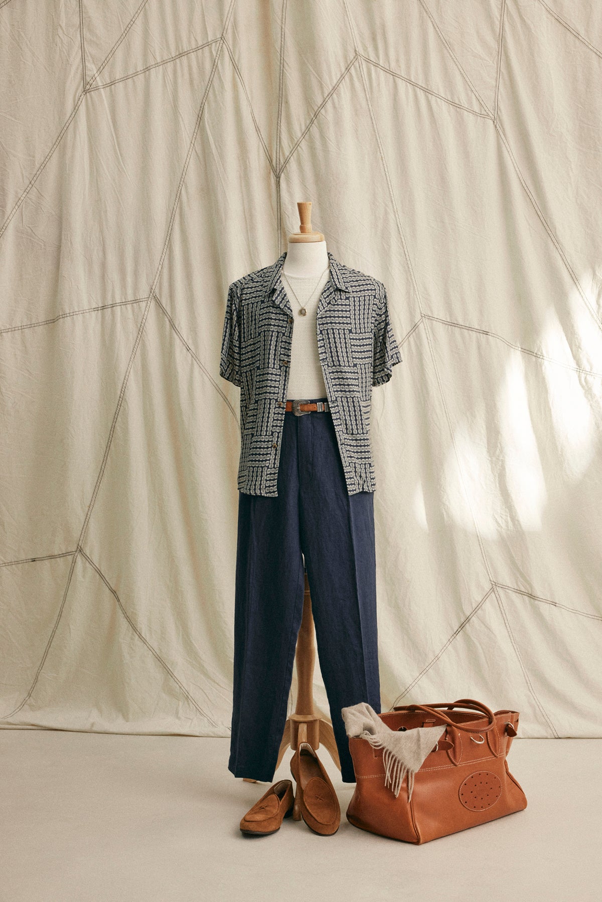 A mannequin in a relaxed fit EMILIO BUTTON-UP SHIRT by Velvet by Graham & Spencer, white tee, navy pants, brown belt and loafers stands beside a large brown leather bag with a scarf.-39152688824513