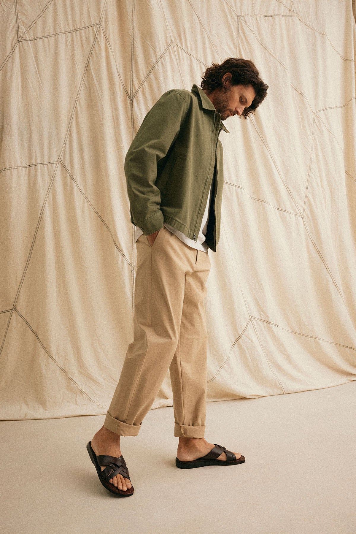 A man in Velvet by Graham & Spencer's STONE PANT stands on a light fabric backdrop, wearing an olive jacket, white shirt, and black sandals while looking down.