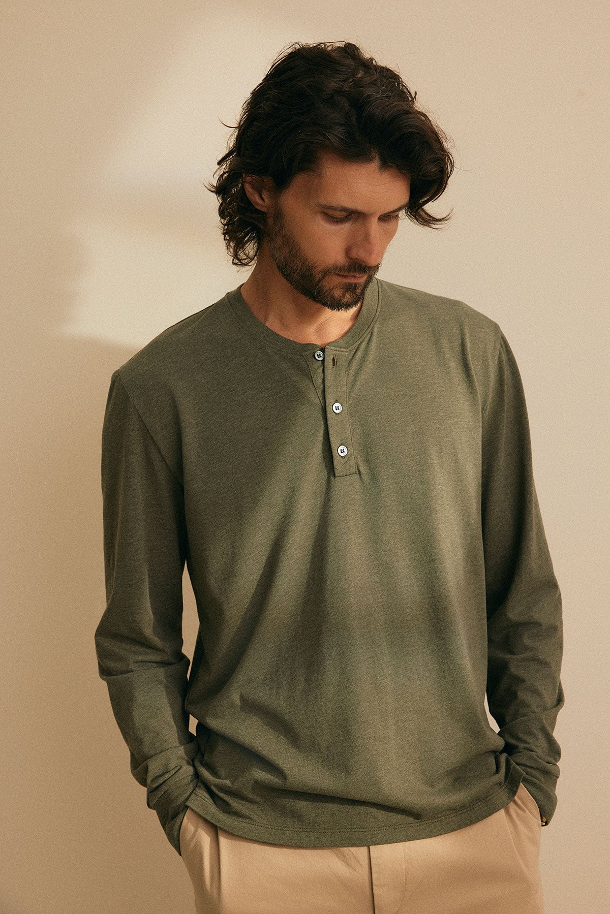 A man with long hair, wearing the DESI HENLEY by Velvet by Graham & Spencer in green, paired with beige pants and a comfortable fit, stands looking down with his hands in his pockets.