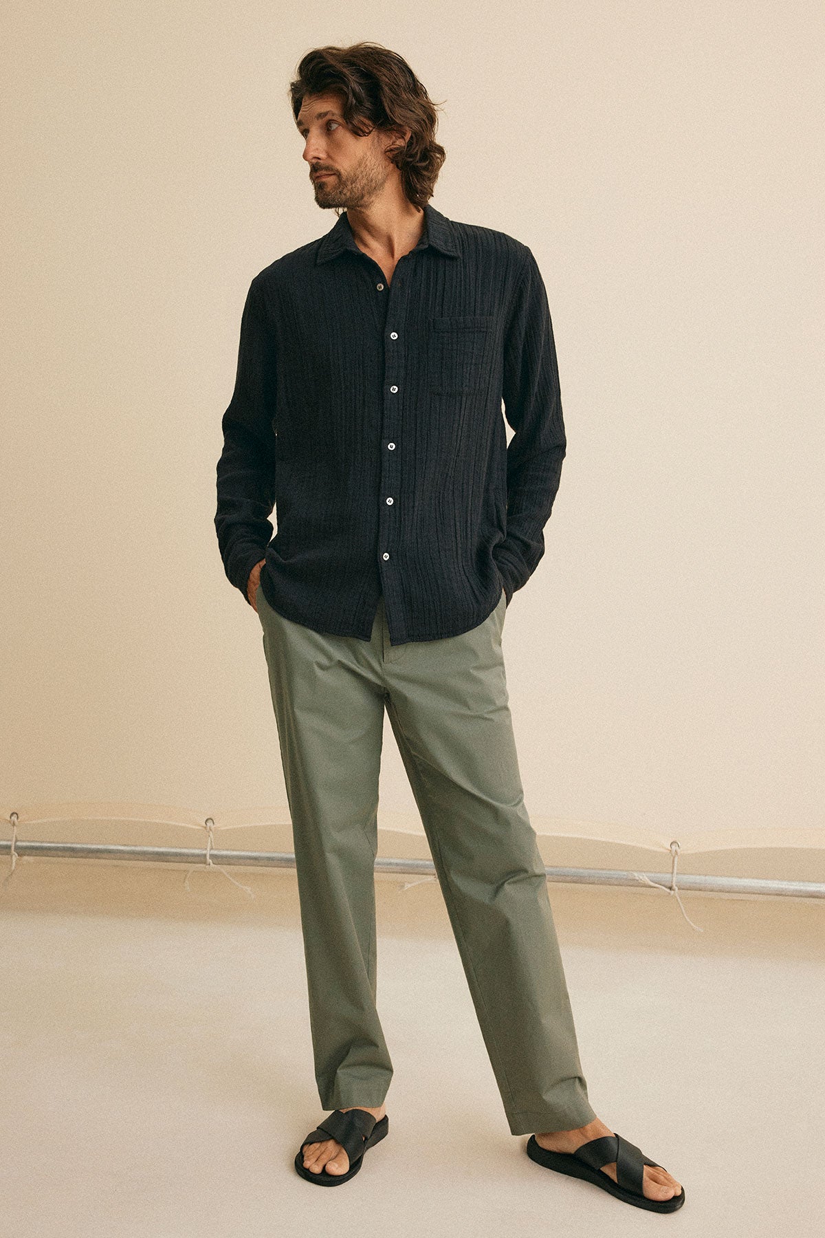 Against a beige background, an individual wears the ELTON COTTON GAUZE BUTTON-UP SHIRT in black by Velvet by Graham & Spencer, complemented with olive green pants and black sandals.