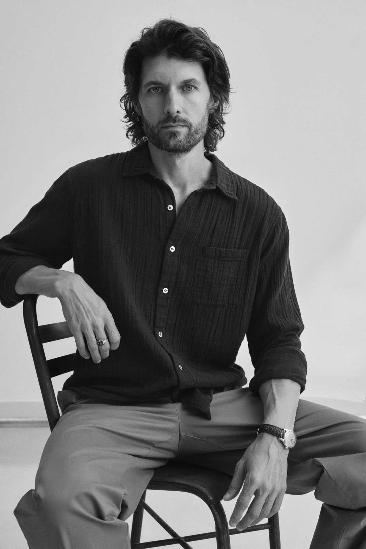 A man with wavy hair and a beard sits on a chair wearing the versatile ELTON COTTON GAUZE BUTTON-UP SHIRT by Velvet by Graham & Spencer, paired with trousers, and looks directly at the camera in this classic black and white photo.