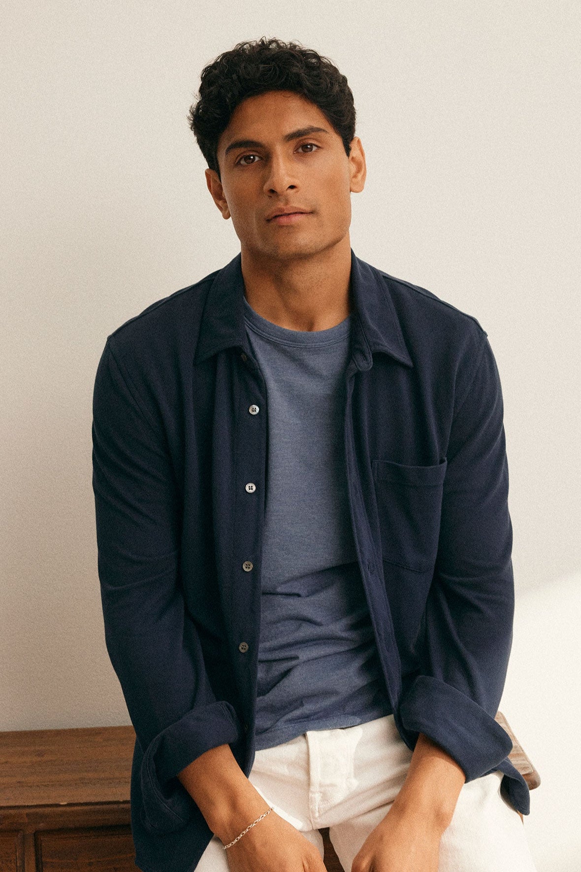 A person with short curly hair sits on a wooden surface, wearing a GIORGIO PIQUE BUTTON-UP SHIRT by Velvet by Graham & Spencer over a T-shirt and light-colored pants, looking at the camera.