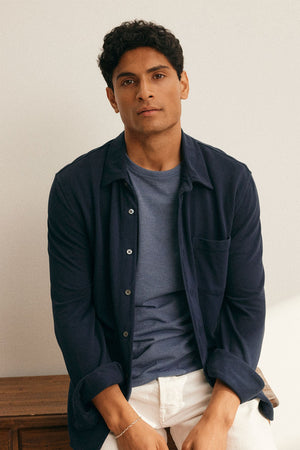 A person with short curly hair sits on a wooden surface, wearing a GIORGIO PIQUE BUTTON-UP SHIRT by Velvet by Graham & Spencer over a T-shirt and light-colored pants, looking at the camera.