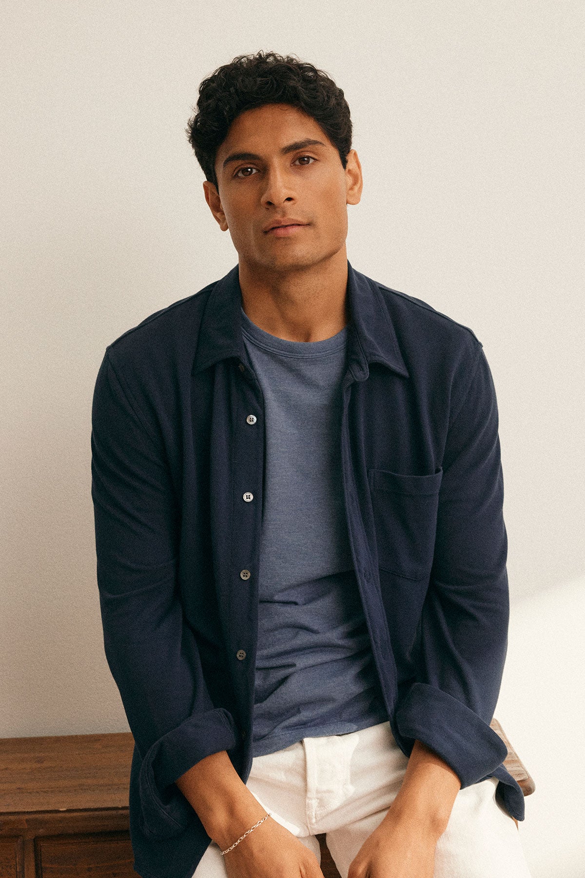 A man in a navy ZANDER TEE by Velvet by Graham & Spencer, offering a relaxed fit, sits on a wooden surface against a light background.