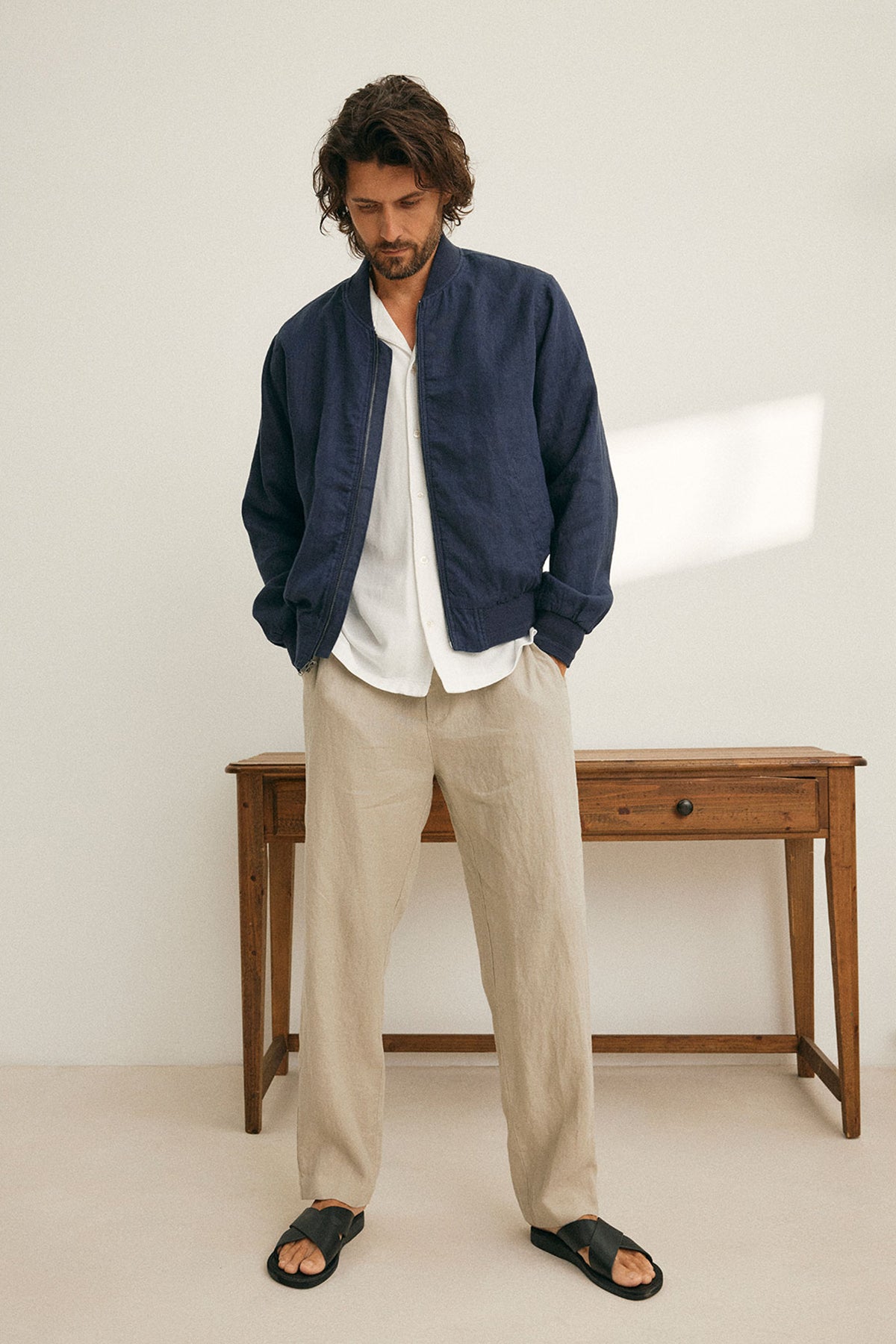 A man in a MURPHY LINEN BOMBER JACKET by Velvet by Graham & Spencer, paired with a white shirt, beige pants, and sandals stands in front of a wooden table, looking down.