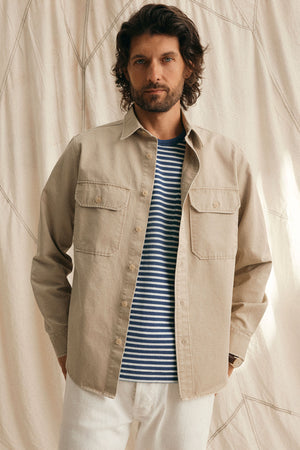 A man with wavy hair wears a beige ORLANDO BUTTON-UP SHIRT by Velvet by Graham & Spencer featuring chest flap pockets over a blue and white striped T-shirt, standing against a neutral fabric backdrop.
