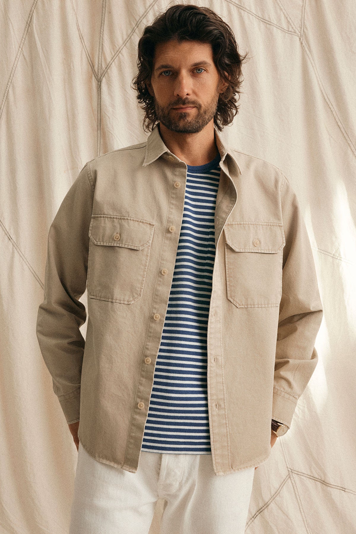 A man with wavy hair stands against a light-colored draped background, wearing a beige button-up shirt over the RIO THERMAL STRIPED CREW by Velvet by Graham & Spencer—a true wardrobe staple.-38877188096193