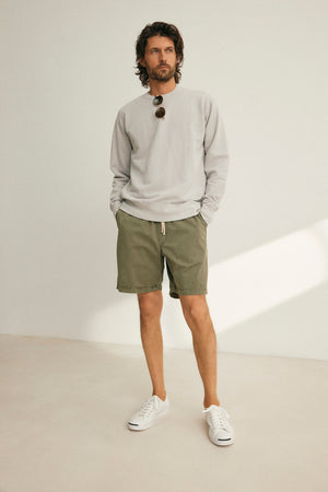 A man stands indoors wearing a light gray sweatshirt, Velvet by Graham & Spencer FIELDER SHORT green shorts with an elastic drawstring waist, white sneakers, and sunglasses hanging from his shirt.