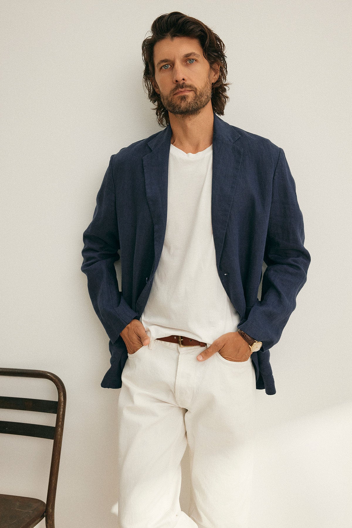 A man with medium-length hair stands against a white wall near a chair, wearing Velvet by Graham & Spencer's WARREN LINEN BLAZER over a white shirt and pants, showcasing classic design.