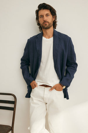 A man with medium-length hair stands against a white wall near a chair, wearing Velvet by Graham & Spencer's WARREN LINEN BLAZER over a white shirt and pants, showcasing classic design.