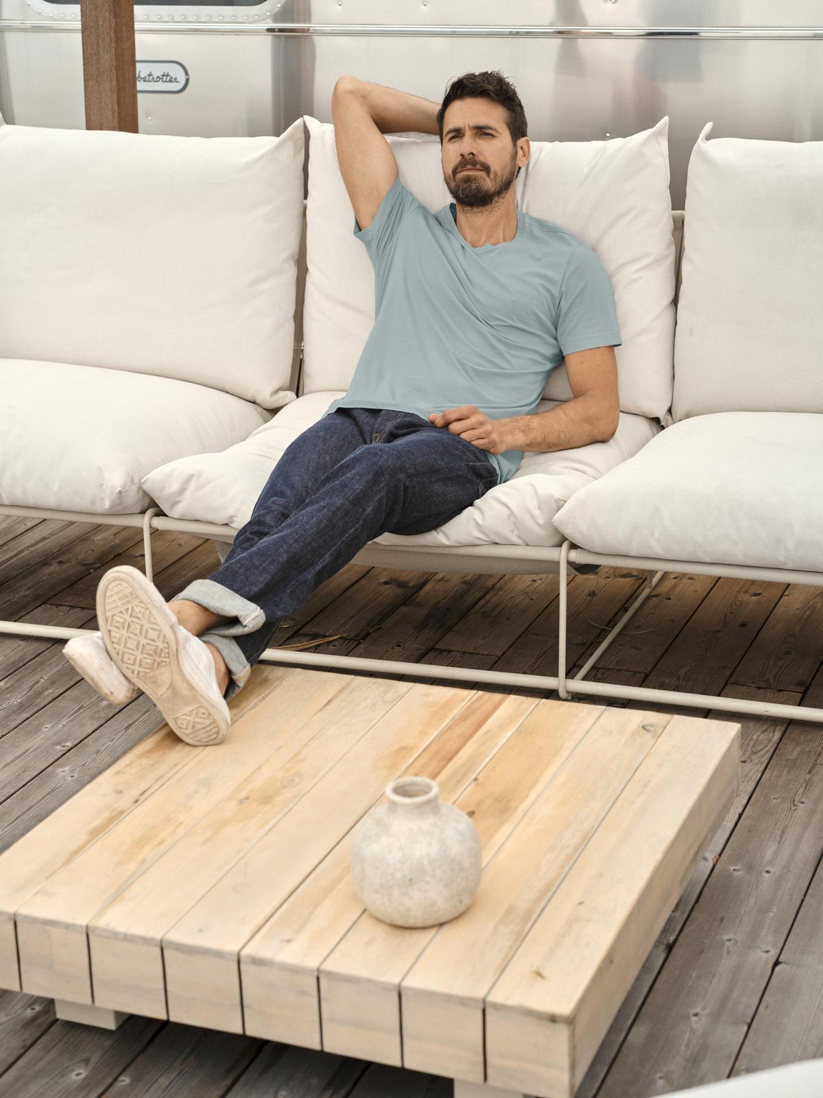 A man is sitting on a white couch, enjoying the vintage-feel softness of the Velvet by Graham & Spencer HOWARD WHISPER CLASSIC CREW NECK TEE.-35782849331393