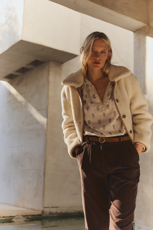 A woman stands indoors wearing the AMARA SHERPA REVERSIBLE JACKET by Velvet by Graham & Spencer over a patterned blouse and brown trousers, with her hands in her pockets.