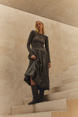 A person with long blonde hair stands on curved concrete steps, wearing the DOLCE DRESS by Velvet by Graham & Spencer and dark boots, set against a beige wall.
