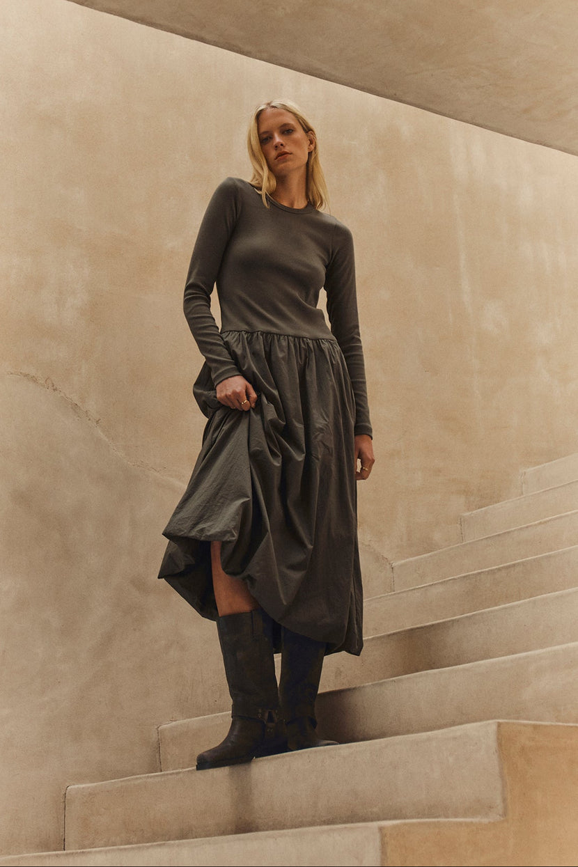 A person with long blonde hair stands on curved concrete steps, wearing the DOLCE DRESS by Velvet by Graham & Spencer and dark boots, set against a beige wall.