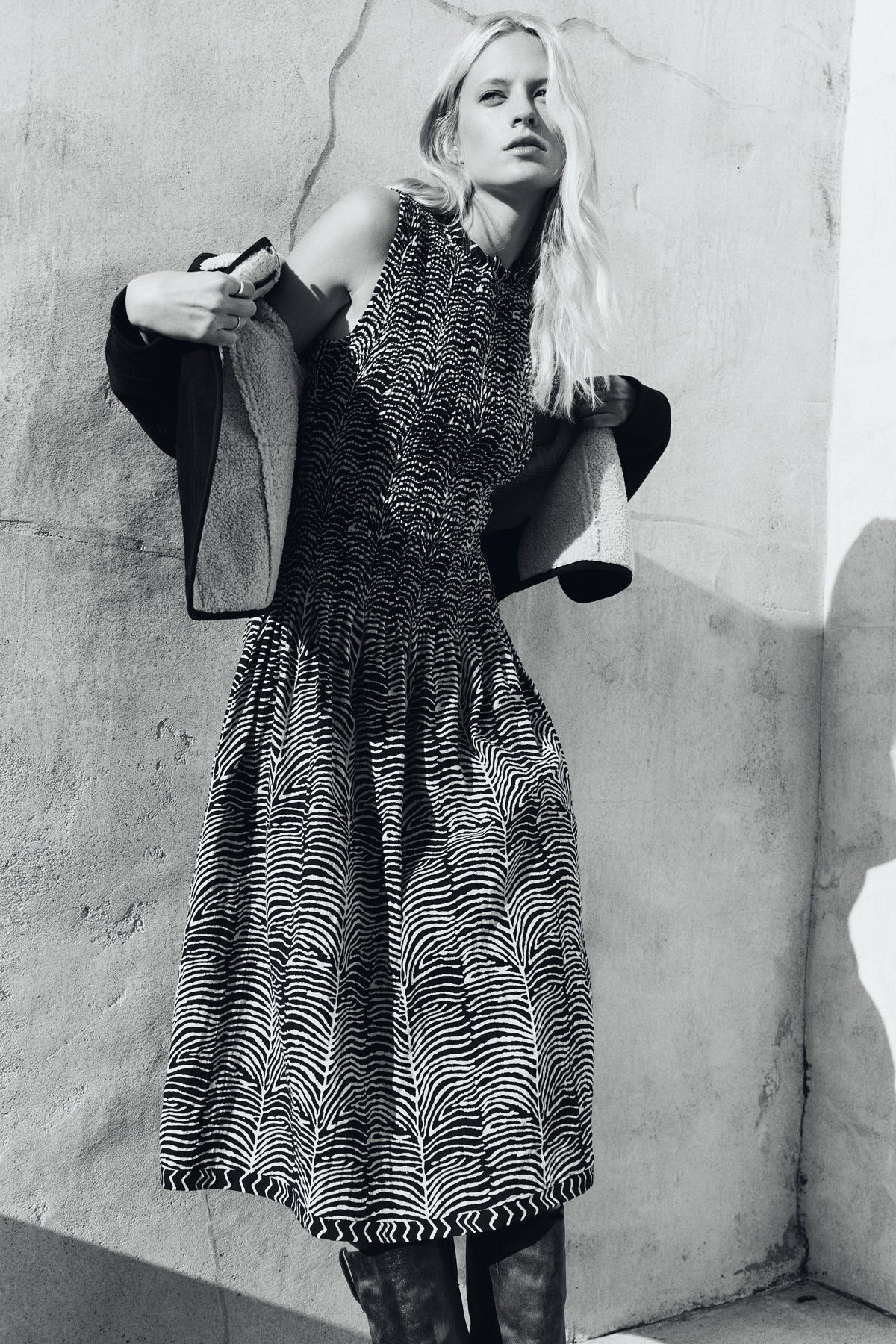 A person stands against a textured wall, wearing the sleeveless, patterned KATHERINE DRESS by Velvet by Graham & Spencer and holding a coat over their shoulders. The image is in black and white.
