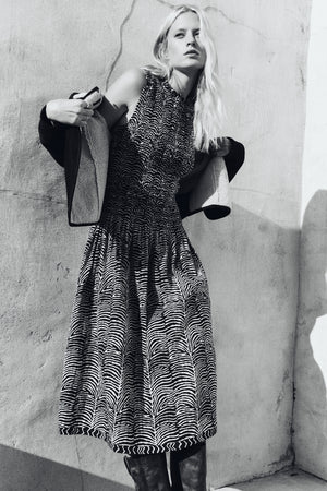 A person stands against a textured wall, wearing the sleeveless, patterned KATHERINE DRESS by Velvet by Graham & Spencer and holding a coat over their shoulders. The image is in black and white.