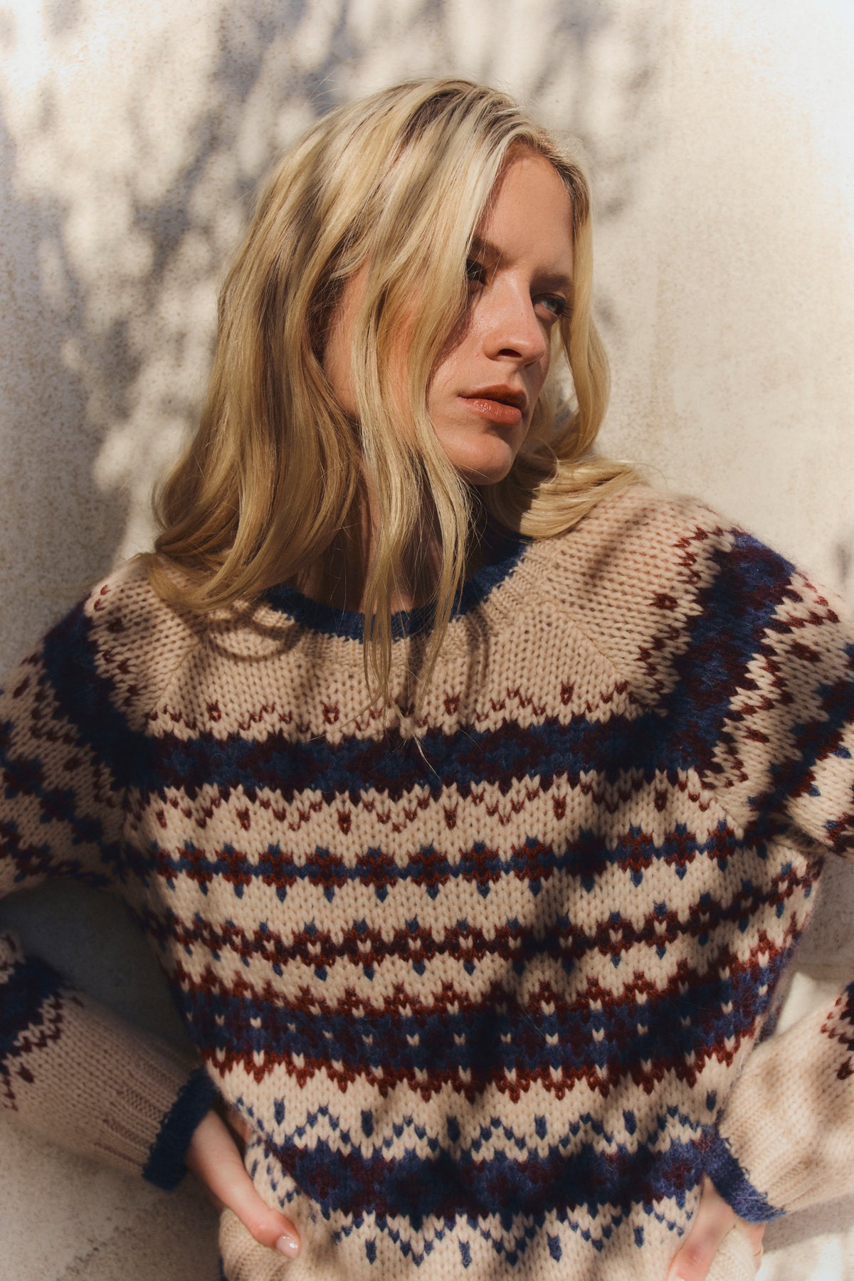 A woman with long blonde hair wears the MAKENA ALPACA SWEATER by Velvet by Graham & Spencer, standing against a light wall with dappled shadows.-39814907527361