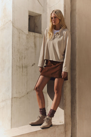 A woman stands against a concrete wall wearing the NILLI POLO long-sleeve top by Velvet by Graham & Spencer, paired with a brown skirt with a side slit, maroon socks, and beige ankle boots.