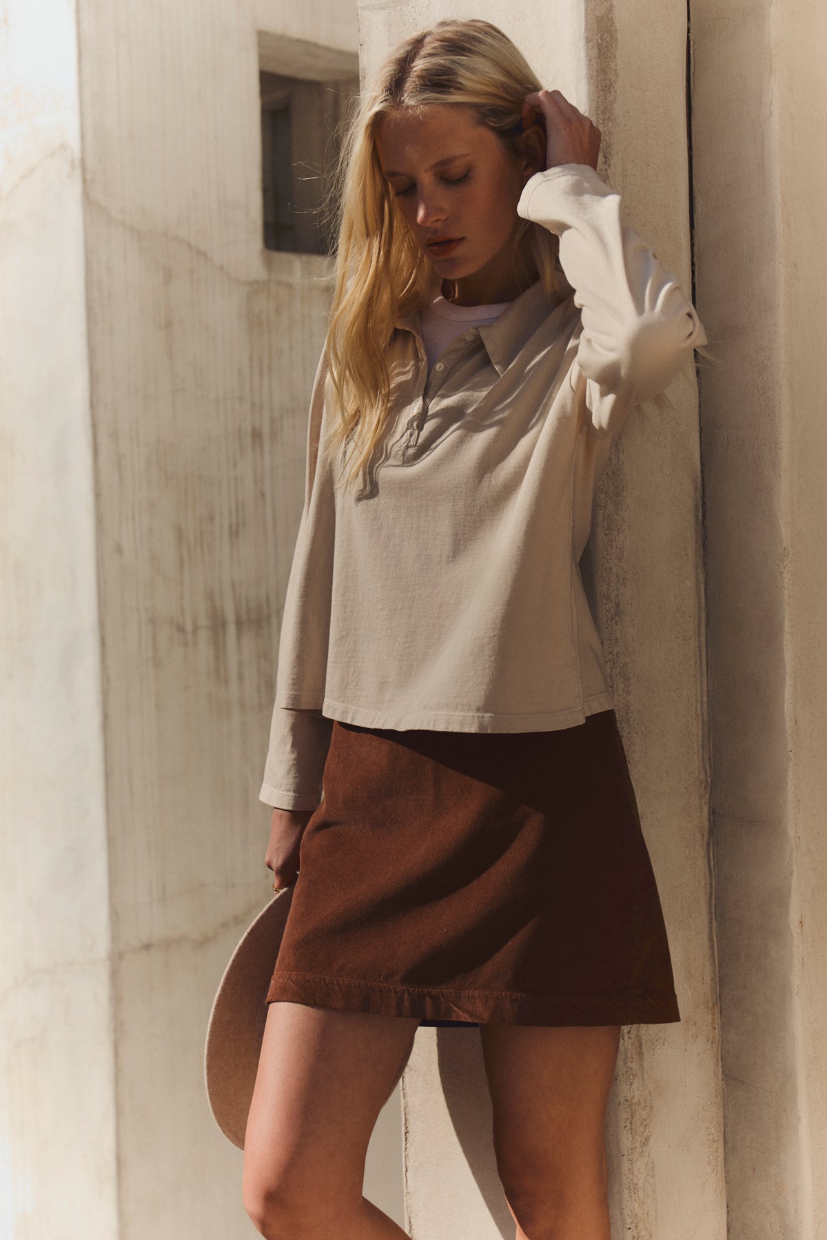 A woman stands by a light wall wearing a beige long-sleeve top and the LIZA CORDUROY SKIRT by Velvet by Graham & Spencer, featuring a fitted silhouette, holding a hat at her side.