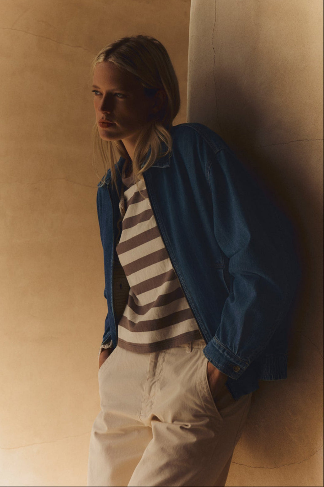 A person stands against a beige wall with hands in pockets, wearing the ANAIS TEE by Velvet by Graham & Spencer under a blue denim jacket and light pants, looking to the side.-39538874777793