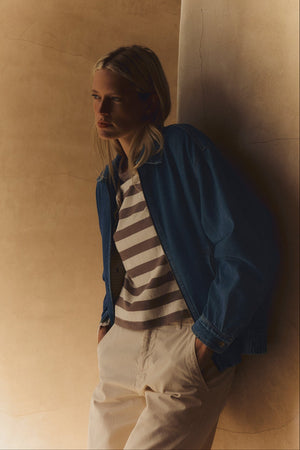 A person stands against a beige wall with hands in pockets, wearing the ANAIS TEE by Velvet by Graham & Spencer under a blue denim jacket and light pants, looking to the side.