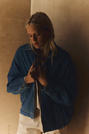 A person with long blonde hair stands against a beige wall, wearing the Velvet by Graham & Spencer PORTER DENIM ZIP-UP JACKET with an elastic hem and light pants, looking down with hands near their collar.