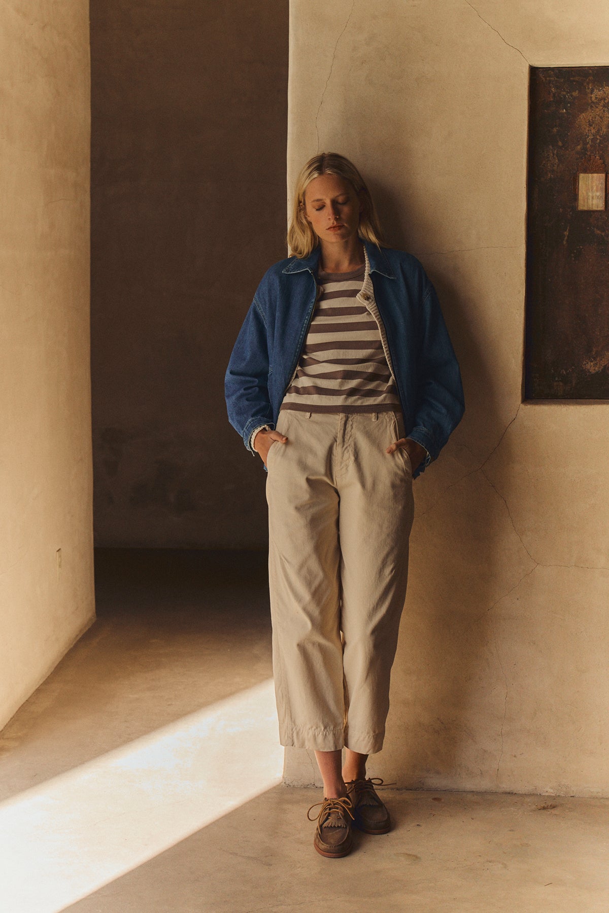 A person stands indoors against a beige wall, eyes closed and hands in pockets, wearing the Velvet by Graham & Spencer PORTER DENIM ZIP-UP JACKET over a striped shirt, with light pants and brown shoes.