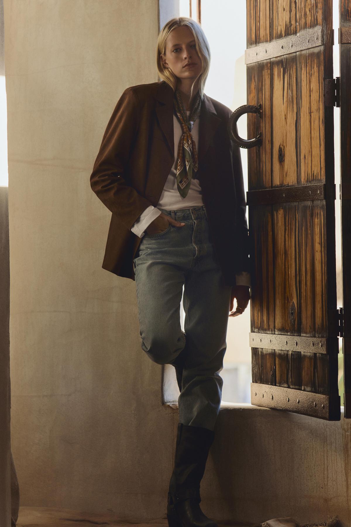 Wearing the SALIE FAUX SUEDE JACKET by Velvet by Graham & Spencer over a white shirt and patterned scarf with blue jeans and black boots, this person stands by an open wooden door in a softly lit room—a timeless layering piece for any occasion.